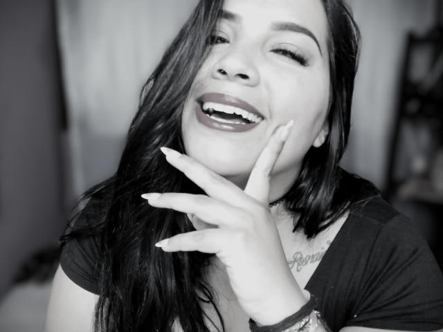 AvaChanne mit dunklem Haar, lächelnd, die ihre glänzenden Lippen zur Schau stellt und mit ihren gepflegten Nägeln auf ihr Kinn zeigt. Sie trägt eine Halskette und hat ein Tattoo auf ihrer Brust..