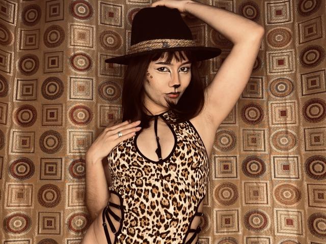 DeisyWest wearing a leopard print dress with cutouts, black strappy sides, and a black hat with a brown band. She has a mole on her right cheek.