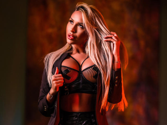 Selena Voss met blond haar, die een zwarte kousenbroek beha draagt, haar grote borsten onthullend, en een open jas, terwijl ze haar haar vasthoudt, verleidelijk kijkend, met oranje verlichting op haar gebruinde huid..