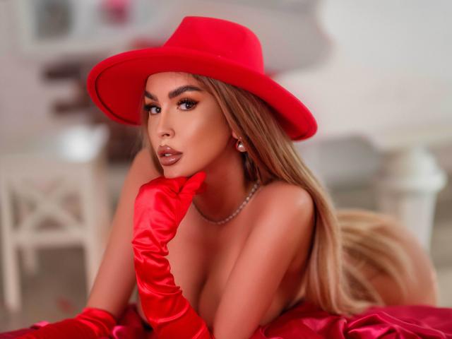 SelenaVoss with long brown hair, wearing a bright red hat, peeks over a gray barrier, showing her brown eyes and arched eyebrows, looking seductive.