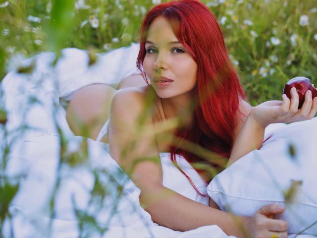 LinaHi with red hair, lying on white pillows, holding a red apple, looking at the camera, showing her tits and ass, in a grassy field with white flowers.
