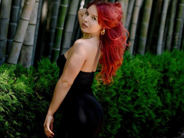 LinaHi with red hair, black strapless dress, gold necklace, standing sideways, showing her ass, green bush in background, bamboo stalks, looking back, hand in hair, sexy.