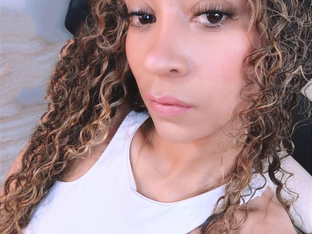 KylieJen with curly brown and blonde hair, wearing a white tank top, has light brown skin and full pink lips, looking slightly to the side with a neutral expression.