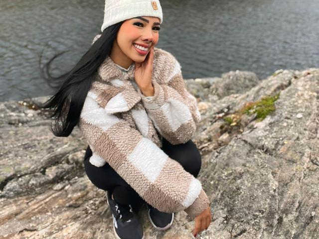 MilaQueen1 with long black hair, wearing a white beanie and a fluffy beige-and-white checkered jacket, squatting on rocky ground, smiling, touching her cheek, wearing black pants and sneakers.