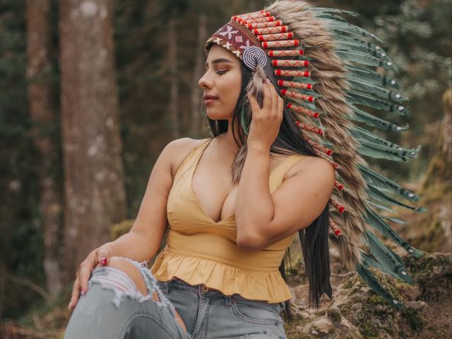 MilaQueen1 with big tits, wearing a yellow top, ripped jeans, and a Native American-style headdress with feathers, touching her face, standing in a forest.