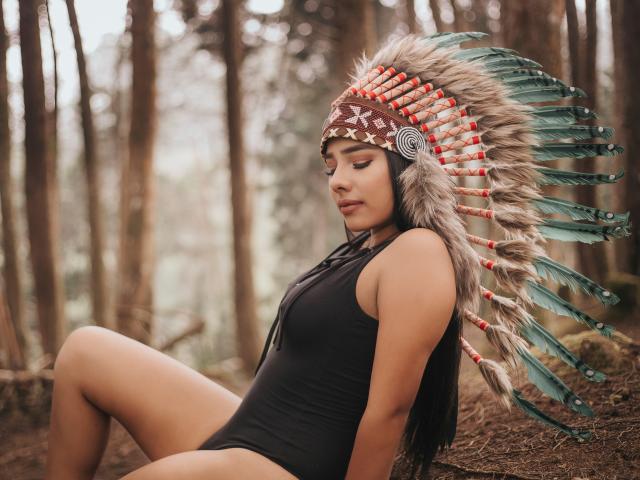 MilaQueen1 with tan skin, long black hair, wearing a black one-piece, sitting in a forest, has a large feather headdress with green and red feathers, and a brown headband.
