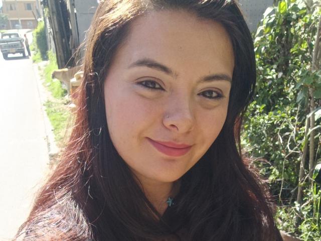 katherinLatin has medium-length brown hair, a nose piercing, and a slight smile with pink lips. She's standing outside with a green vine on the right.