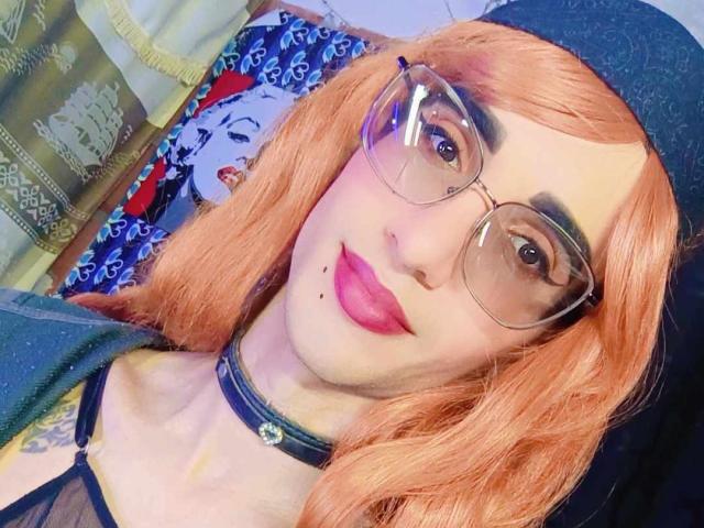 KrissiriWat with orange hair, glasses, black choker, and pink lipstick, wearing a black cap, showing a tattoo on the left shoulder, and a small mole on the chin.