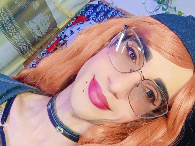 KrissiriWat with orange hair, glasses, and pink lipstick, wearing a choker, smiling at the camera, showing a beauty mark on her left cheek. Boobs not visible.
