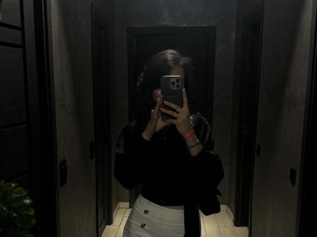 LillianSunny en una habitación oscura, selfie frente al espejo, top negro, pantalones blancos de cintura alta, chaqueta negra, sosteniendo el teléfono, cubriéndose la cara, pulsera rosa, cabello castaño oscuro, de pie, fondo oscuro..