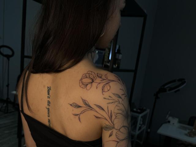 LillianSunny's upper back and shoulder, featuring a floral tattoo and a cursive tattoo on her right shoulder blade, with dark brown hair, in dim lighting.
