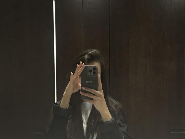 Lillian Sunny, con cabello oscuro, vistiendo una chaqueta negra, sosteniendo un iPhone plateado, cubriéndose la cara con ambas manos, en una habitación oscura con paneles de madera..