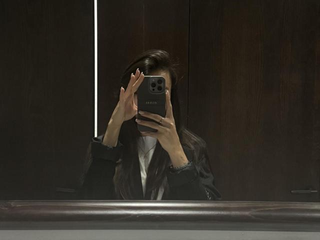 LillianSunny taking a mirror selfie with her iPhone, hands covering her face, wearing a black jacket over a white shirt, dark hair partially covering her eyes.