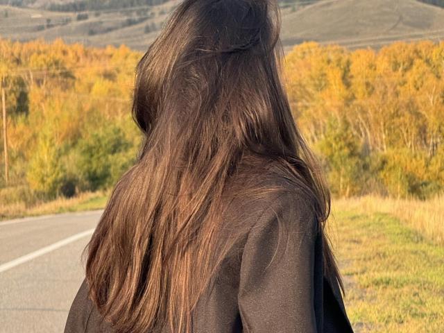 El cabello largo y castaño de LillianSunny le cubre la espalda mientras camina por un camino, vistiendo una camisa negra, con árboles otoñales y colinas en el fondo..