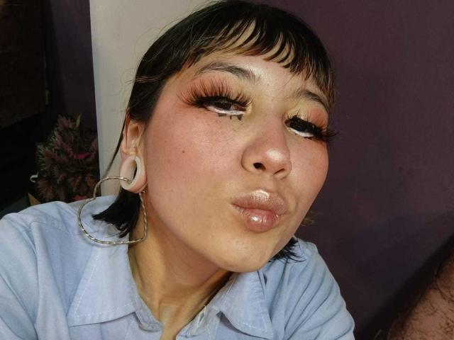 LalaKlein with puffy glossy lips, wearing large silver hoop earrings, has dark hair with bangs, and is making a pouty face, dressed in a light blue shirt.
