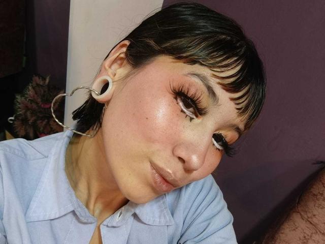 LalaKlein mit kurzen, schwarzen Haaren, die ein weißes Hemd trägt, silberne Creolen und sichtbare, weiße Ohrpiercings, hat ein süßes Lächeln und strahlende Haut, und hält den Kopf leicht geneigt..