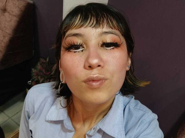 LalaKlein with dark brown hair, pink eyeshadow, and long eyelashes, pouting her full lips in a light blue shirt, wearing large white earrings, in a slightly dim room.
