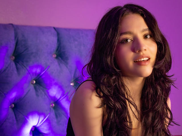 KateCrown with wavy dark brown hair, cute smile, and brown eyes, wearing a black top, has her tits slightly visible, against a blue tufted headboard with purple lighting.