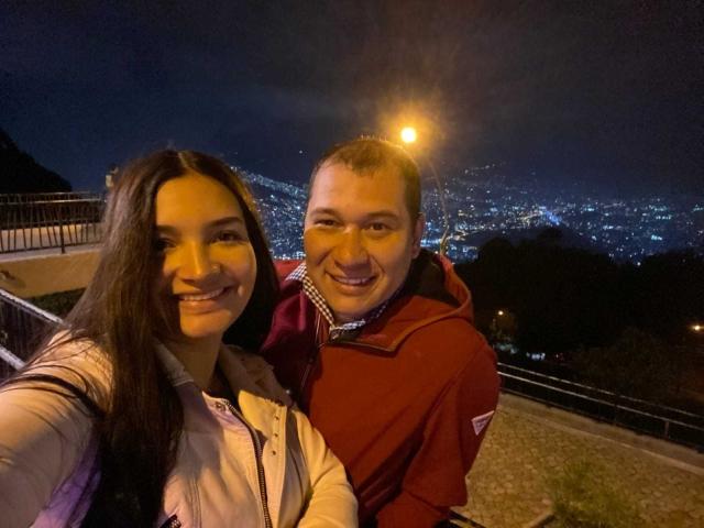 TazFer tiene senos, cabello largo y castaño, y una camisa blanca, de pie junto a TazFer, que tiene la cabeza calva, una sudadera roja y una sonrisa, por la noche con las luces de la ciudad..