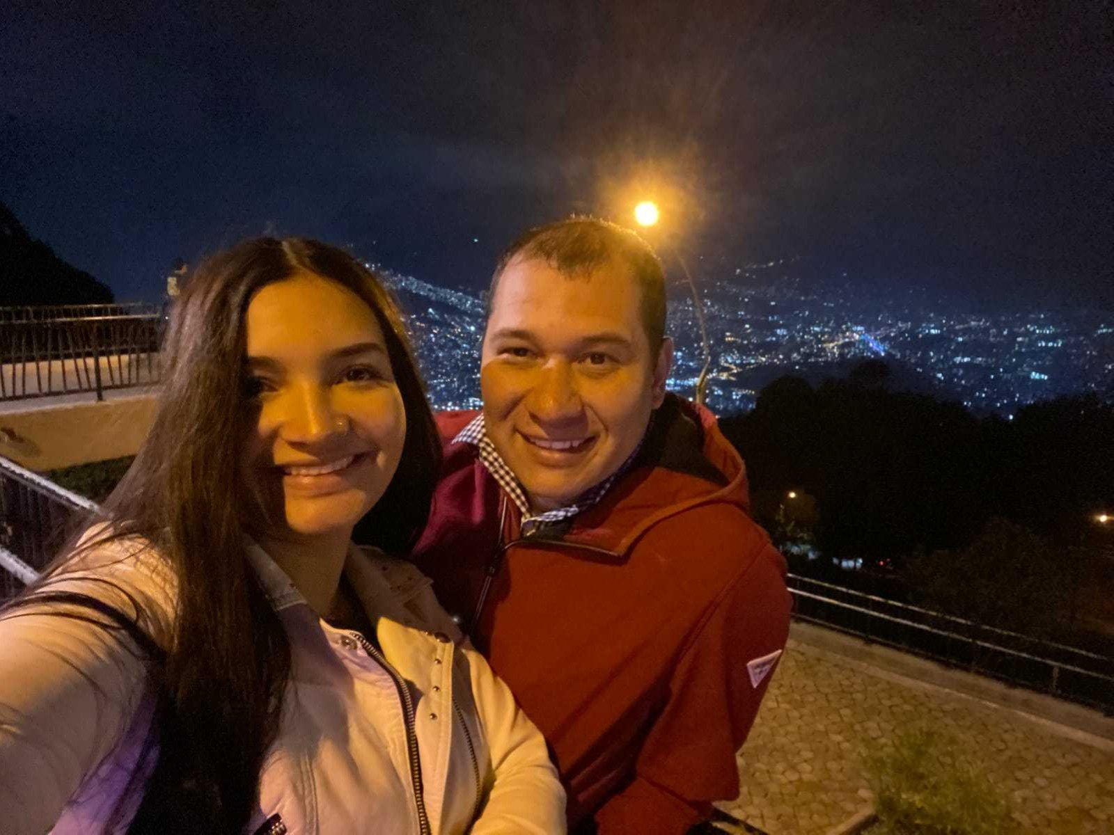 TazFer tiene senos, cabello largo y castaño, y una camisa blanca, de pie junto a TazFer, que tiene la cabeza calva, una sudadera roja y una sonrisa, por la noche con las luces de la ciudad..