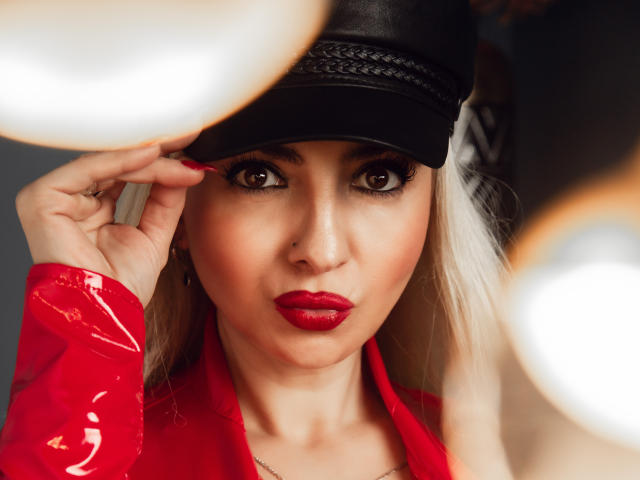 Vixenia with red glossy jacket, black leather cap, and red lipstick, holding cap with red-manicured fingers, staring into camera with intense brown eyes. Blurred white orbs in foreground.