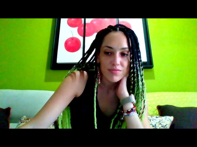 Kailani69 with braided green and black hair, leaning on a couch, wearing a black top, wrist bracelet, and watch, with a green wall and red balloon artwork behind.