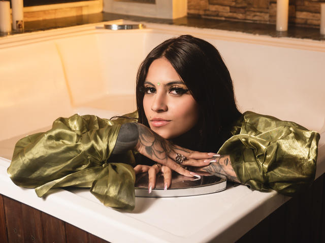CoriPaige, con cabello oscuro, un vestido de satén verde, manos tatuadas y maquillaje dorado, apoyada en una bañera blanca, mirando a la cámara, mostrando sus labios carnosos y uñas brillantes..