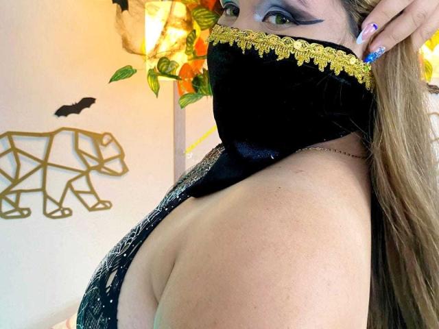 SalmaAzaman with black velvet mask, gold lace trim, and blue eyeshadow, showing her tits in a sparkly black dress, blonde hair, and purple nails, in a selfie.