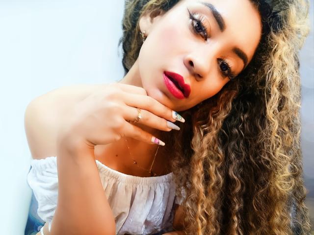 KylieJen with curly blonde hair, red lipstick, and white off-shoulder top, holding her chin, showing manicured nails, gold necklace, and rings, looking seductive.
