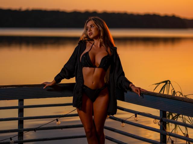 CapriceVonDe com cabelos longos e loiros, um sutiã de renda preto e calcinha preta, vestindo uma camisa preta aberta, apoiada em um corrimão ao pôr do sol, com água e árvores ao fundo..