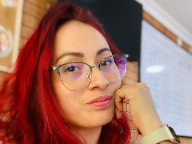 JulietaMartin with red hair, wearing glasses, and a slight smile, resting her chin on her hand, showing her pink lipstick and white wristwatch.