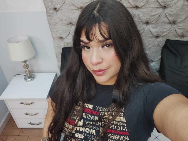AntonelaPark with long dark hair, wearing a black shirt, smiling at the camera, showing a cute face, with pink lipstick, standing next to a white nightstand with a lamp.