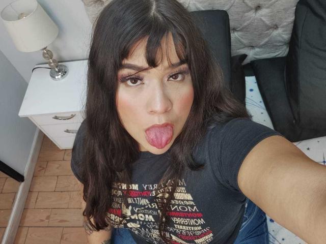 AntonelaPark with long dark hair, brown eyes, and bangs, sticking her tongue out, wearing a black T-shirt and blue jeans, sitting on a bed, taking a selfie.