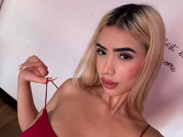 ChristieGarci with blonde hair, red dress, holding a strap, pouty lips, pink blush, and long eyelashes, looking at the camera. Boobs visible, attractive, cute.