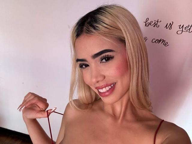 ChristieGarci has blonde hair, dark eyebrows, and is smiling with pink blush on her cheeks. She's holding a red string, showing her tits with a red strap top.