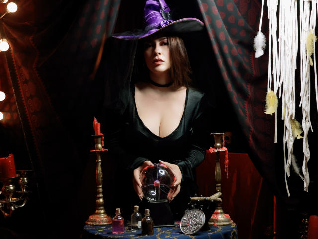 ChristineBray with dark brown hair, black choker, deep cleavage in black dress, purple witch hat, holding a glass mask, red candles, and bottles on a dark table.