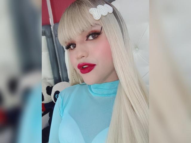 SweetNatashaX with long blonde hair, red lipstick, and blue top, has a white hair clip and large eyes, sitting in a white chair, showing her cute face and tits.