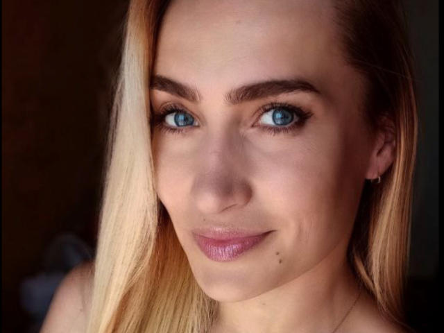 KateOweens has blue eyes, blonde hair, and pinkish lips with a small beauty mark under her right eye. She's smiling softly, looking directly at the camera.