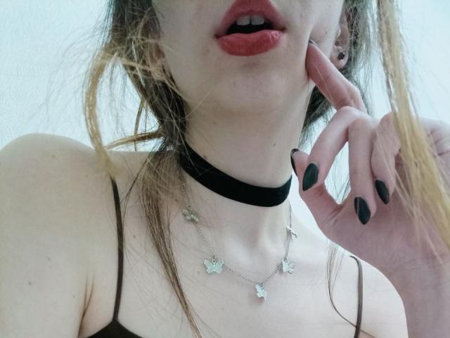 WollyMolly com pele clara, lábios vermelhos, colar preto, colar de borboleta, unhas pretas, tocando o queixo, cabelo castanho e alças finas, mostrando o peito e a boca..