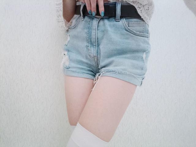Les cuisses pâles de WollyMolly, vêtue de shorts en jean bleu clair usés, avec une ceinture noire, un haut en dentelle et des bas blancs montants jusqu'aux genoux, arborant des ongles turquoise, se dressent devant un mur blanc texturé..