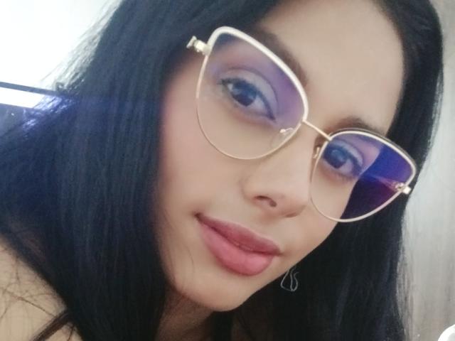 AnaisHilton with long black hair, purple eyeshadow, gold-rimmed glasses, and pink lips, showing a slight smile, wearing small hoop earrings, looking directly at the camera.