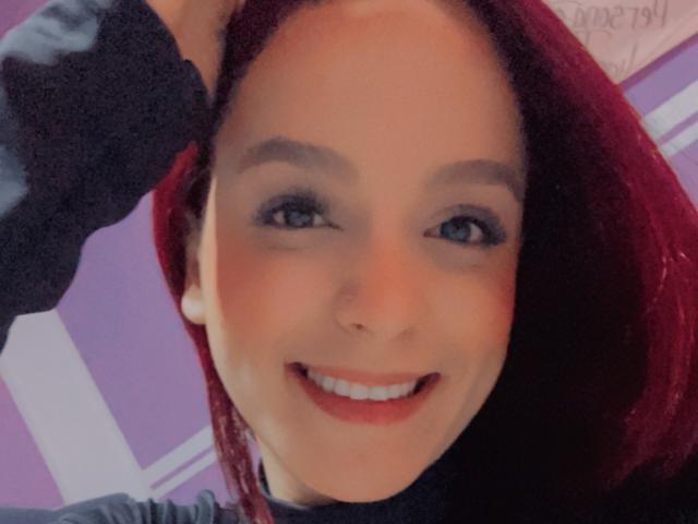 MaiaPark has red hair, blue eyes, and is smiling with red lipstick. She's wearing a black sweater, holding her hair up. Her tits are not visible.