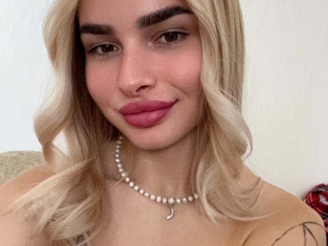 LunaHeadly has blonde wavy hair, pink lipstick, and a pearl necklace with a crescent moon pendant. She's smiling with her tits slightly visible and a tattoo on her arm.