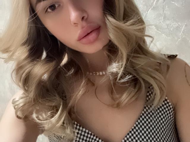 LunaHeadly with blonde, wavy hair, wearing a black-and-white checkered dress, pearl necklace, and pouty pink lips, looking slightly up. Boobs visible, soft lighting, gentle facial expression.