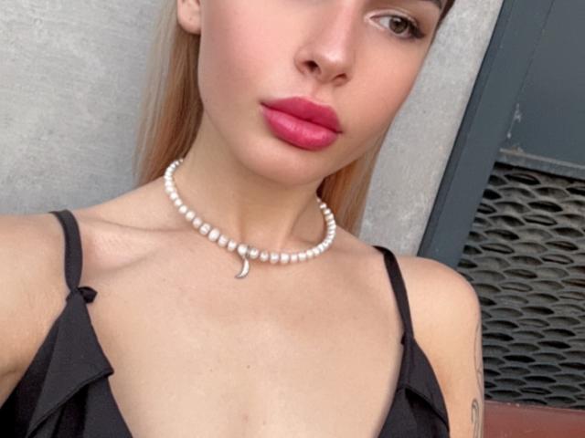 LunaHeadly has light skin, pink lipstick, and a white pearl necklace with a crescent moon charm, wearing a black dress with thin straps, showing her boobs.