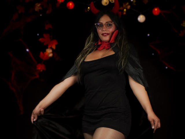 VeronicaSaienz, with large tits, wearing a black dress, red bow, and glasses, has long green-tipped hair, dark cape, and a sexy smile, in a dark, festive setting.