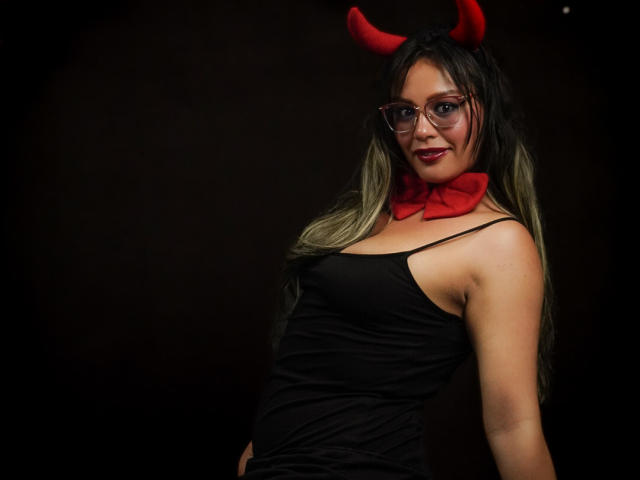 VeronicaSaienz with red devil horns, glasses, and a red bowtie, wearing a black, spaghetti-strap, tight-fitting dress, smiling at the camera. Her tits are small, her ass is cute.