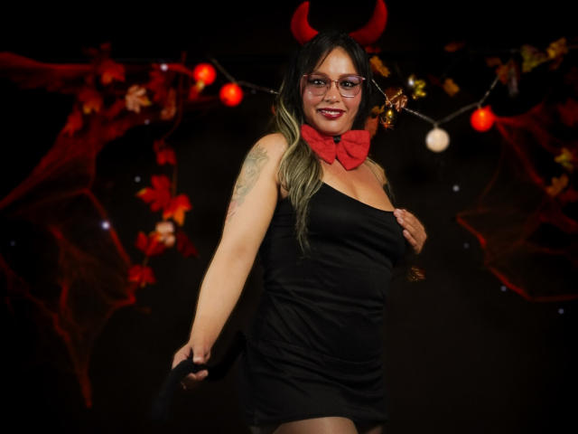 VeronicaSaienz in a black dress, red devil horns, red bowtie, glasses, and holding a black mask, smiling with tattoos on her right arm, in a dark room.