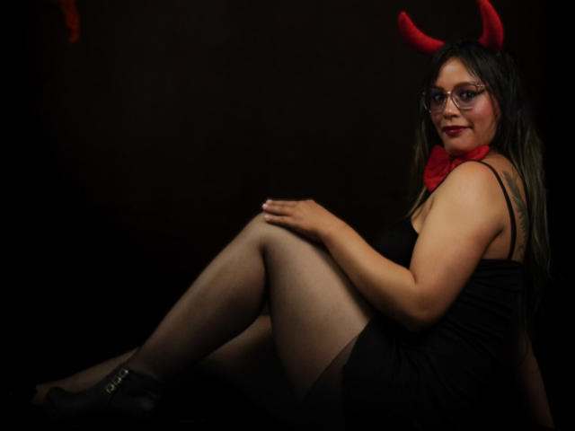 VeronicaSaienz with red devil horns, glasses, black dress, red bowtie, and black stockings, sitting with knees up, showing her ass, smiling, dark background.