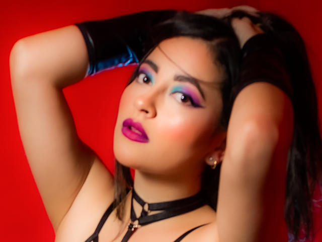 LunaTikka with purple lipstick, blue eyeshadow, and black strappy top, hands in long black hair, wearing black gloves, red background, looking up with seductive eyes.